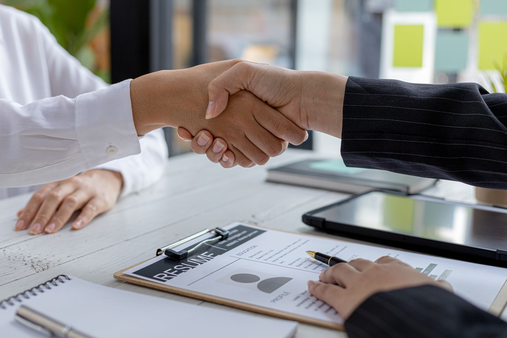 Two people shaking hands over a desk with a resume, notebook, and tablet visible.