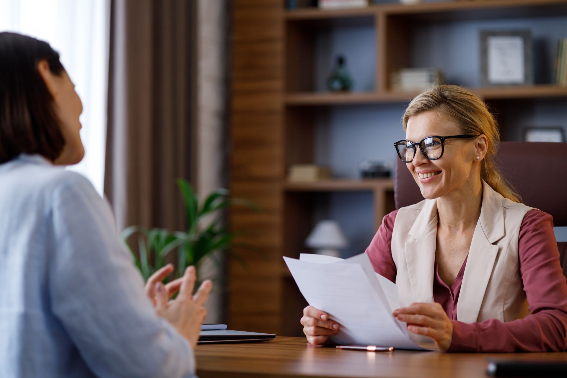 Person with glasses reviewing documents while talking to another person in an office. Person with glasses reviewing documents while talking to another person in an office.