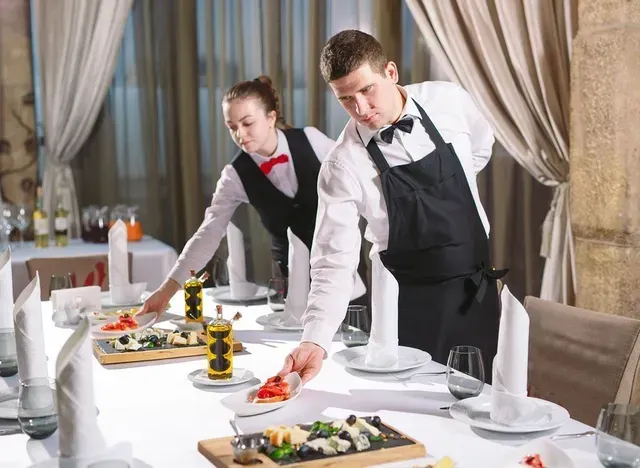Two servers setting a table in a restaurant. One adjusts a plate, the other reaches for food.