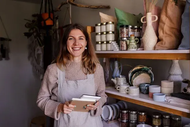 Woman smiling, holding notepad, standing in shop with shelves of handmade goods.