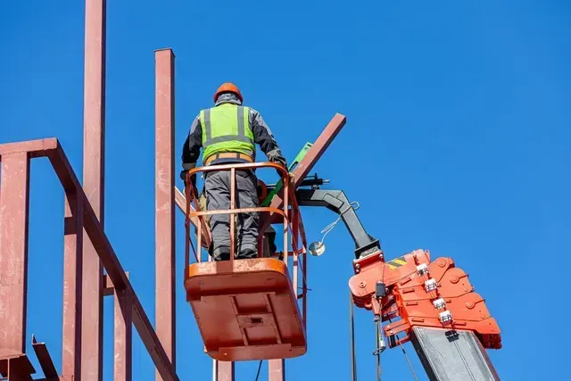 Construction worker in an orange lift platform working on steel structure against a clear blue sky.