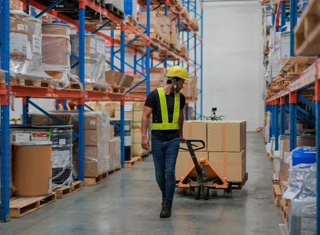 Warehouse worker moving boxes on a pallet jack. Wearing safety vest and hard hat, surrounded by shelves of inventory.