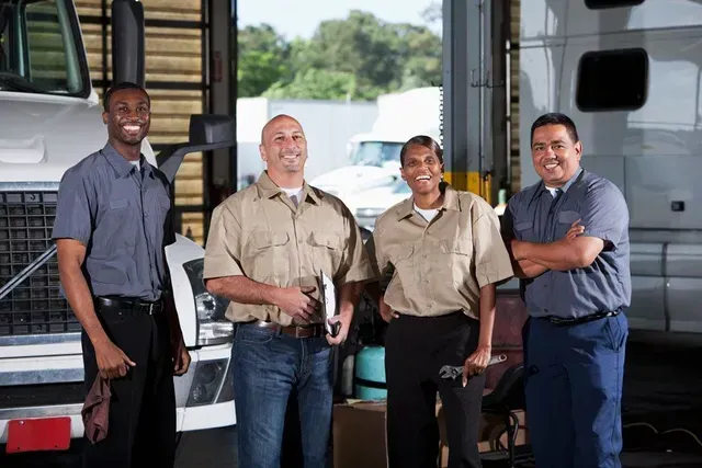 Four people, possibly mechanics, smiling in front of a semi-truck in a garage.