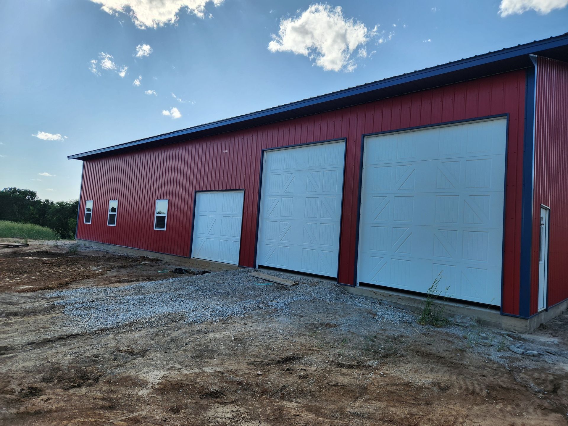 Red metal building with three garage doors, a small window, and gravel in front under a blue sky.