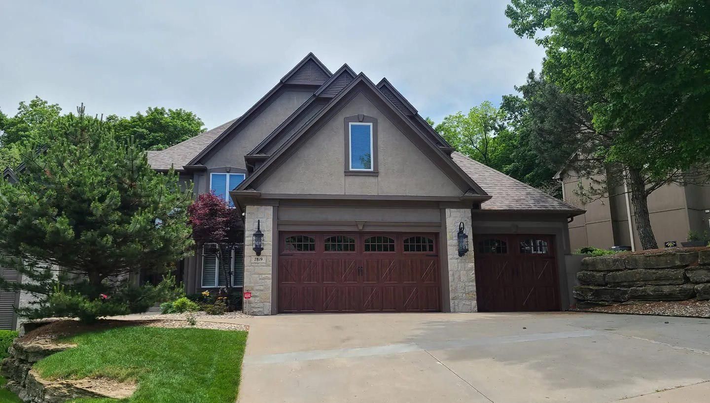 Brown and tan house with a two-car garage, red doors, and a stone facade. Lush green trees frame the home.