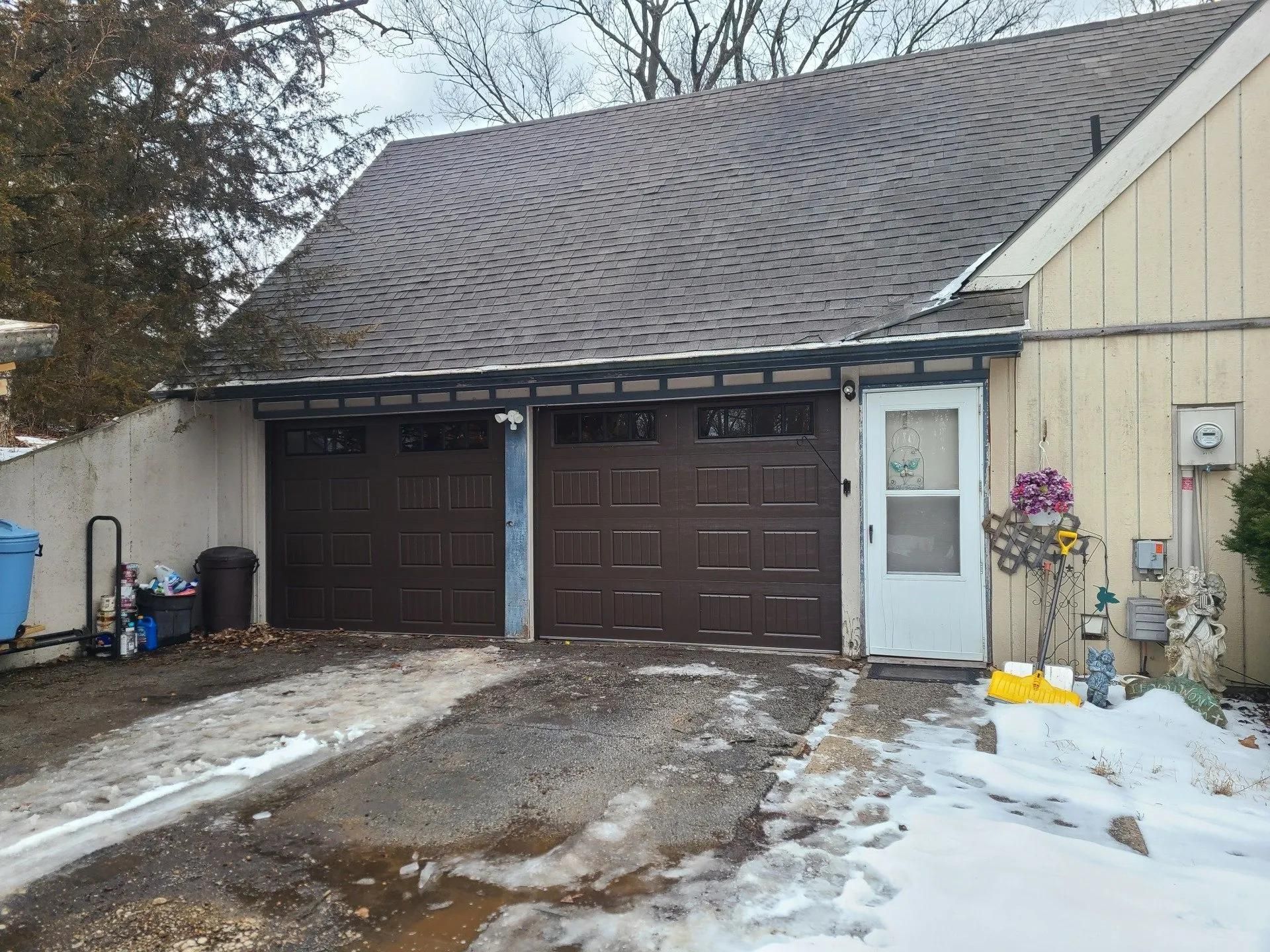 Brown garage doors, white door, snow, ice, asphalt driveway, beige building exterior.