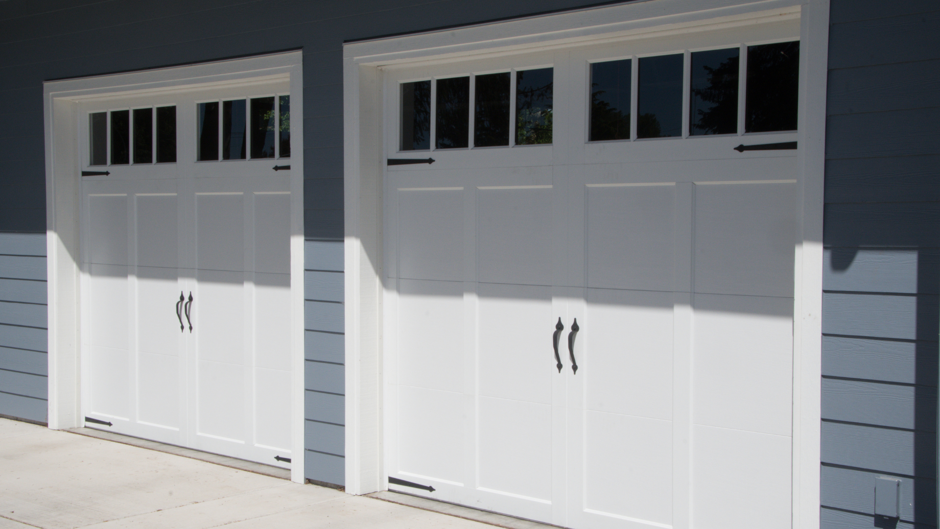 Two white garage doors with black hardware and window panels, set in a light blue exterior.