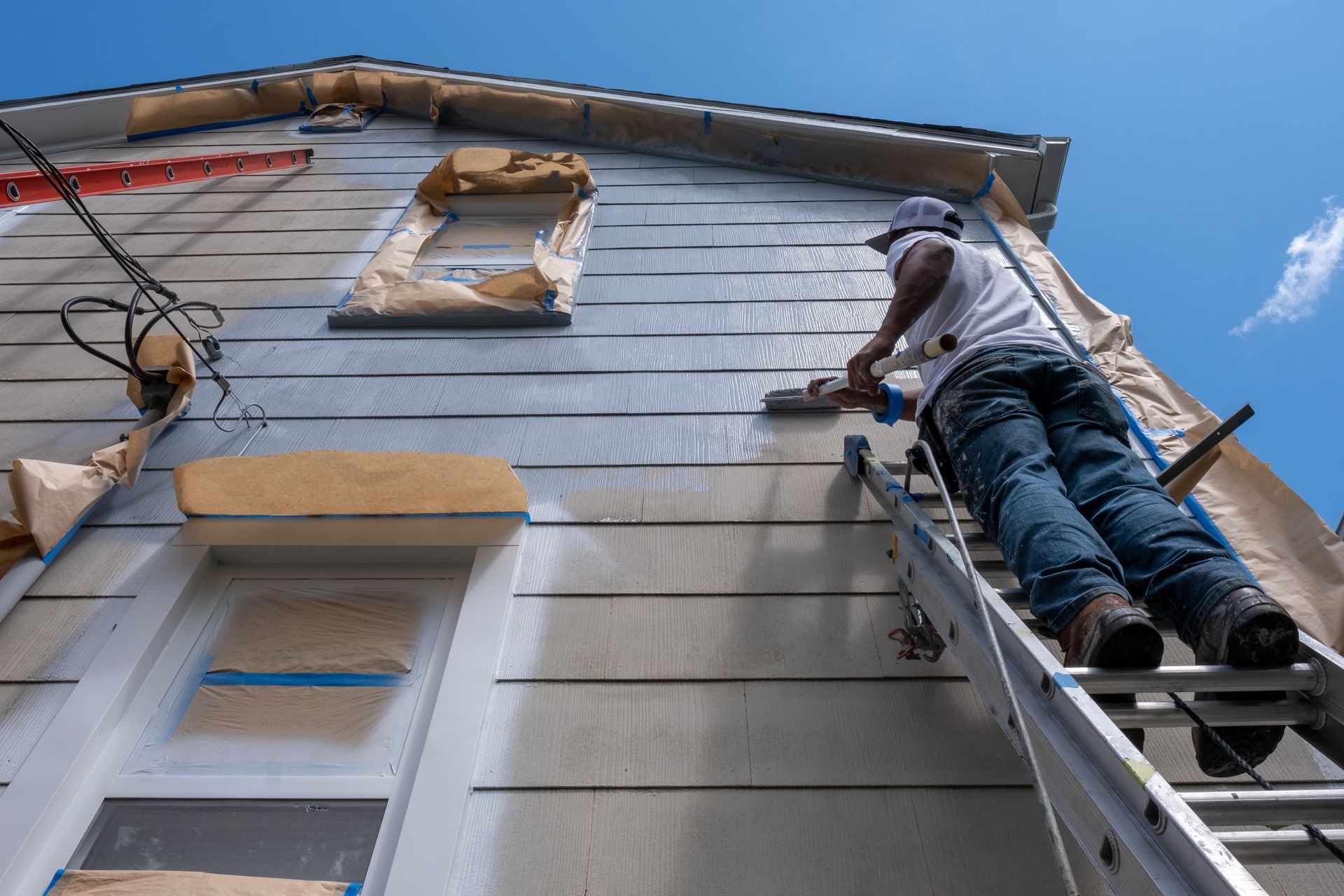 An exterior house painter on a ladder is painting the exterior of a house.