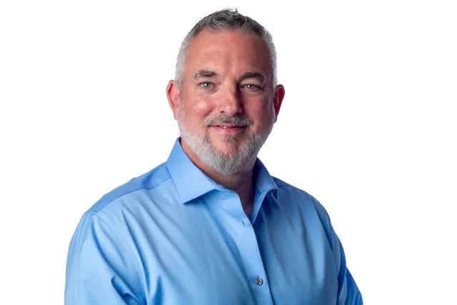 A man with a beard is wearing a blue shirt and smiling.