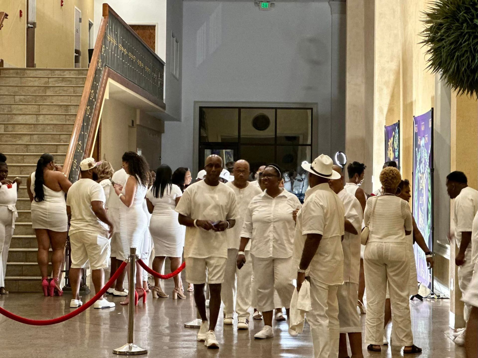 People in white clothing at an indoor event, walking near artwork, a staircase, and a doorway.