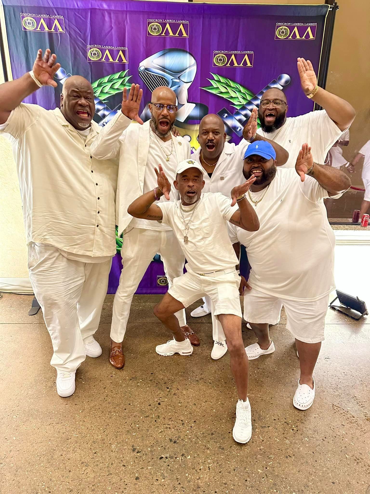 Group of six people in white outfits pose excitedly in front of a banner.