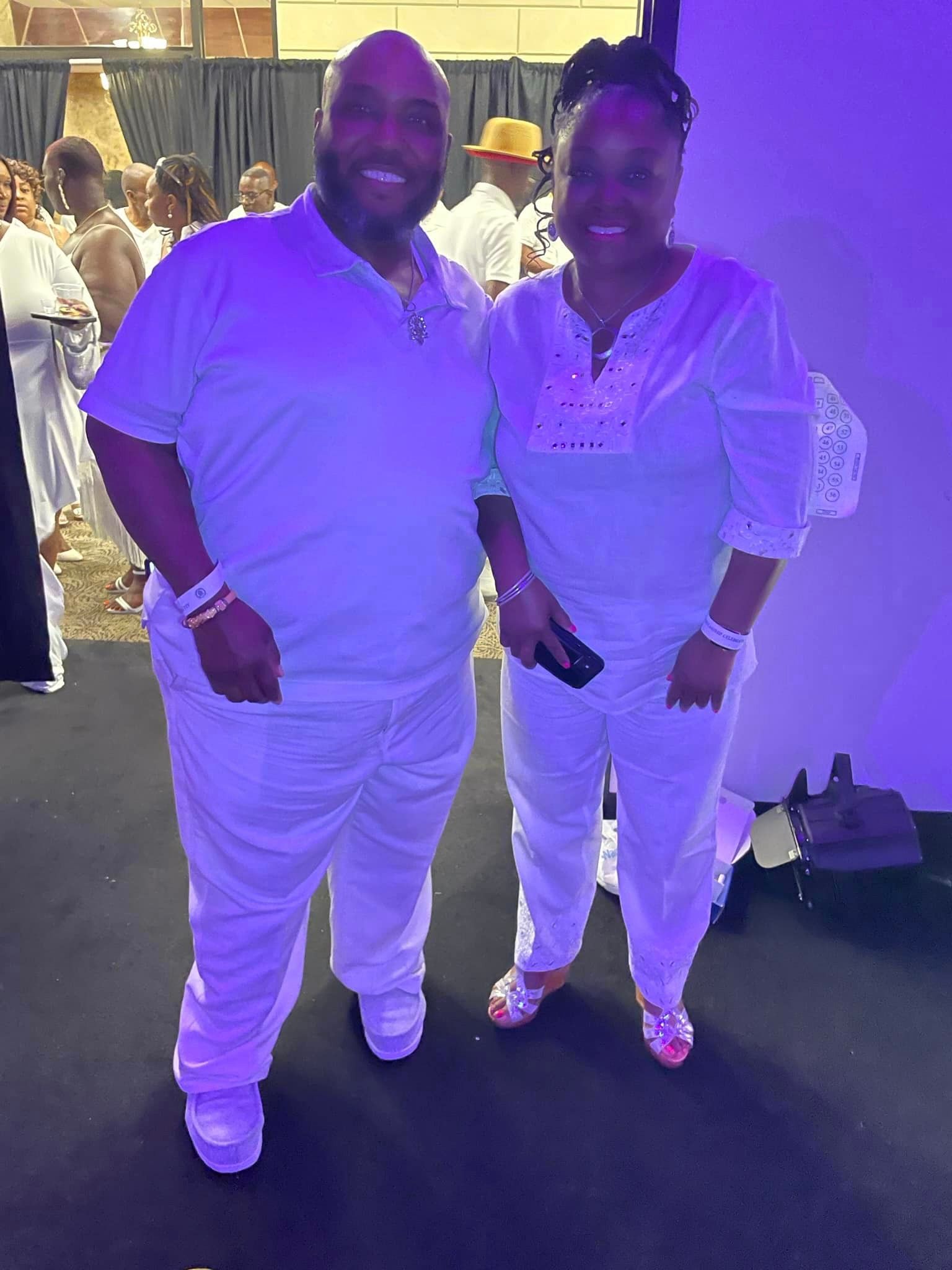 Two people, dressed in white outfits, pose for a photo at an event with purple lighting.