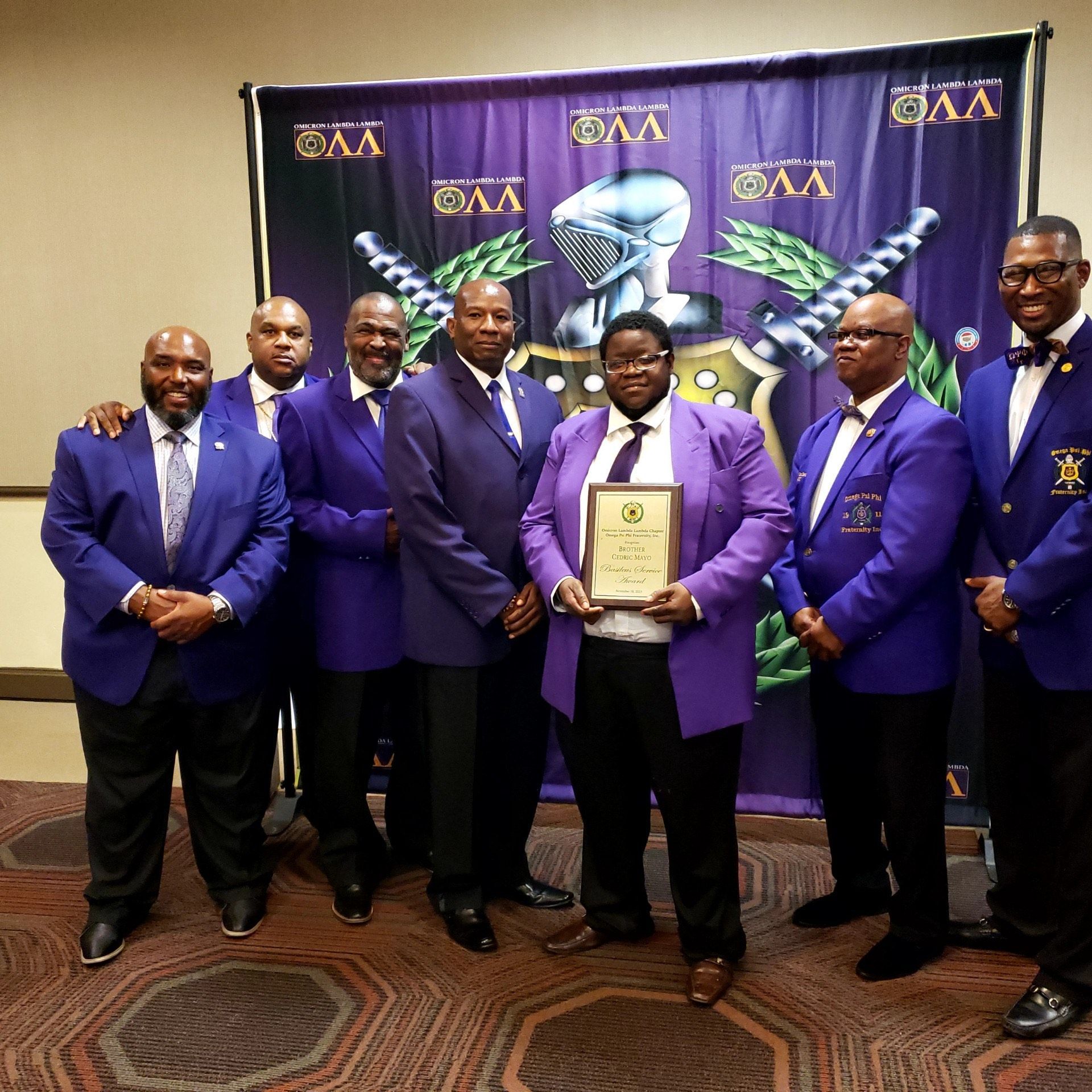 Group of men in purple and blue suits, holding plaque, in front of a banner with a knight emblem.
