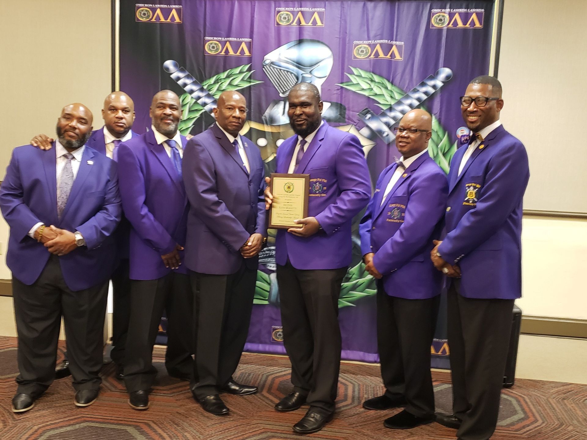 Men in purple suits hold a plaque in front of a banner.