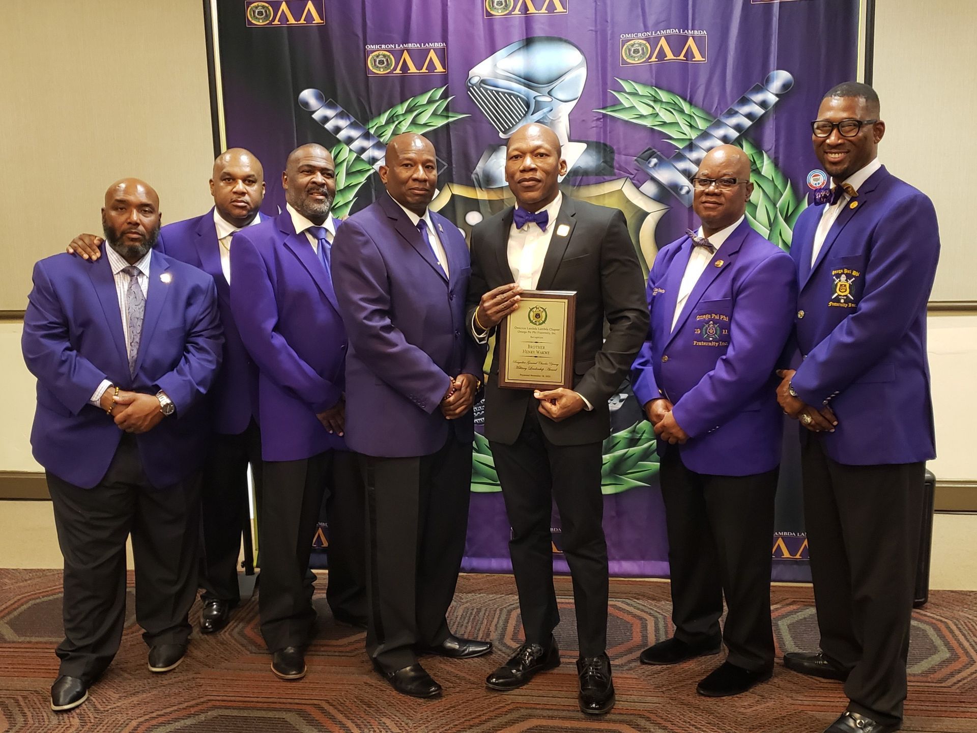 Group of men in suits, some in purple, holding an award in front of a banner.