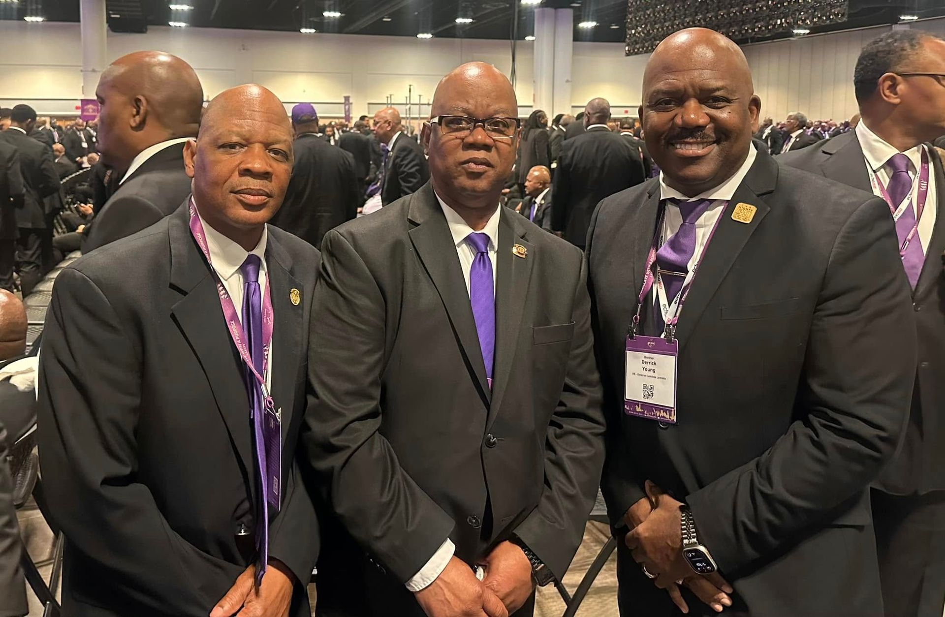 Three men in suits pose, purple accents on lapels and ties, indoors, smiling.