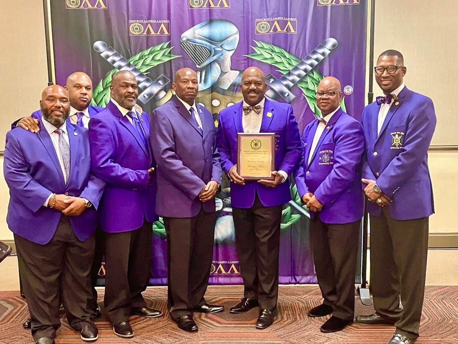 Group of men in purple jackets holding an award, standing in front of a banner.