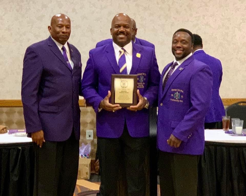 Three men in purple blazers and ties; one holds a plaque. Indoor setting with a table.
