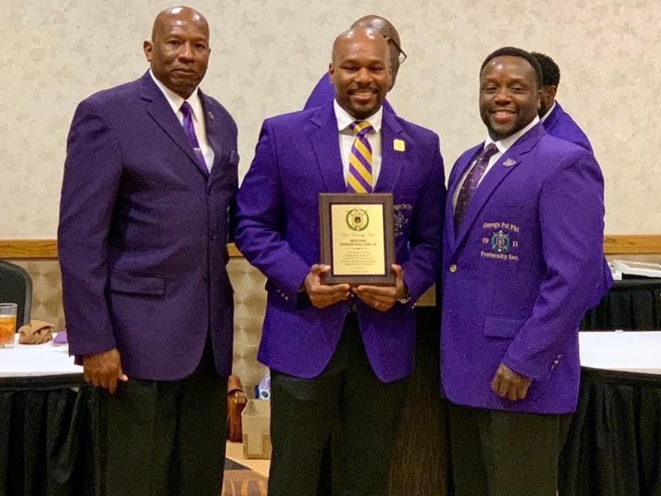 Three men in purple jackets at an event, one holding a plaque, smiling.