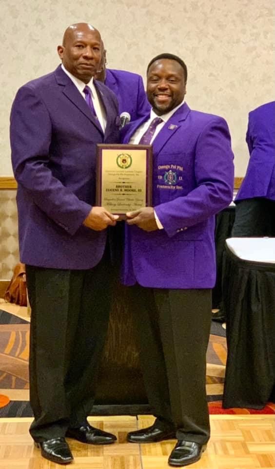 Two men in purple jackets holding a gold plaque, smiling. Indoor setting with a table.