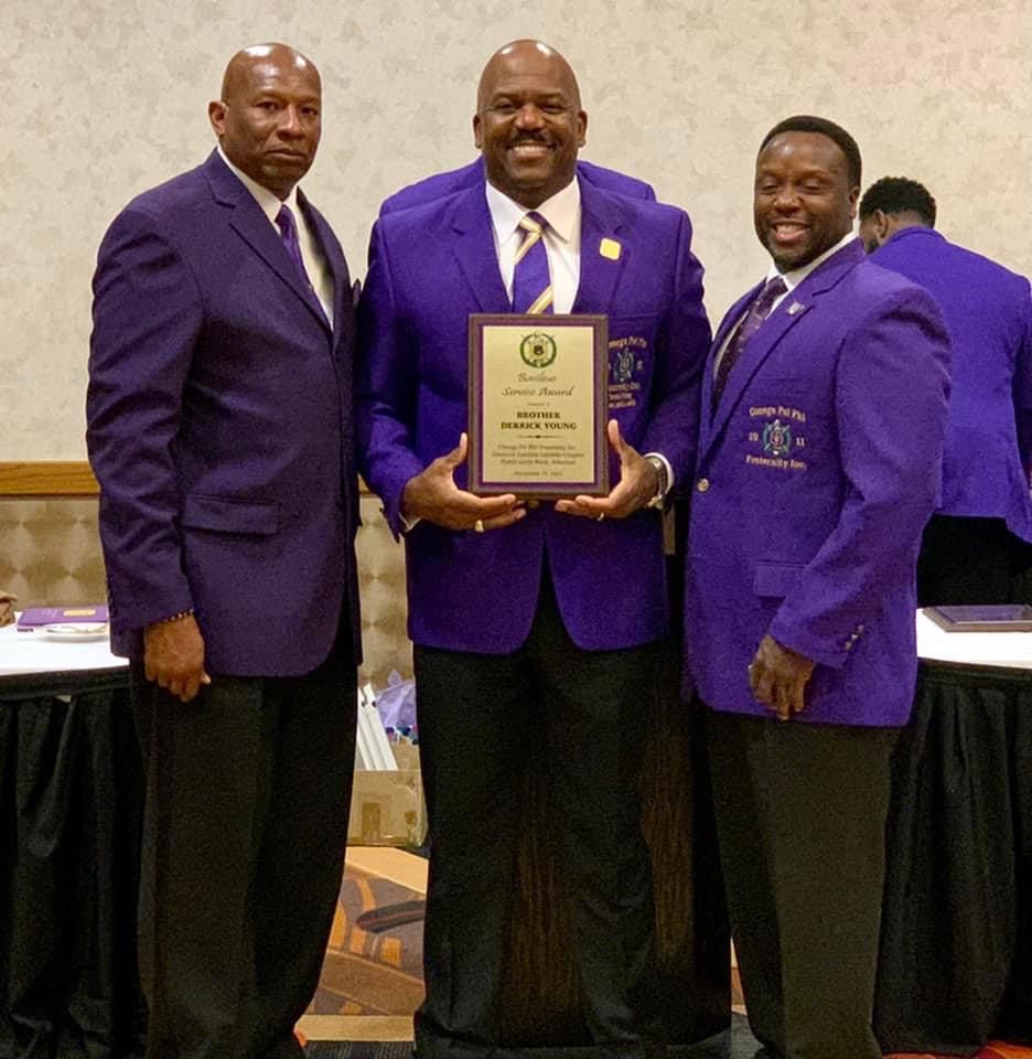 Three men in purple jackets pose with an award, likely at a conference or event.