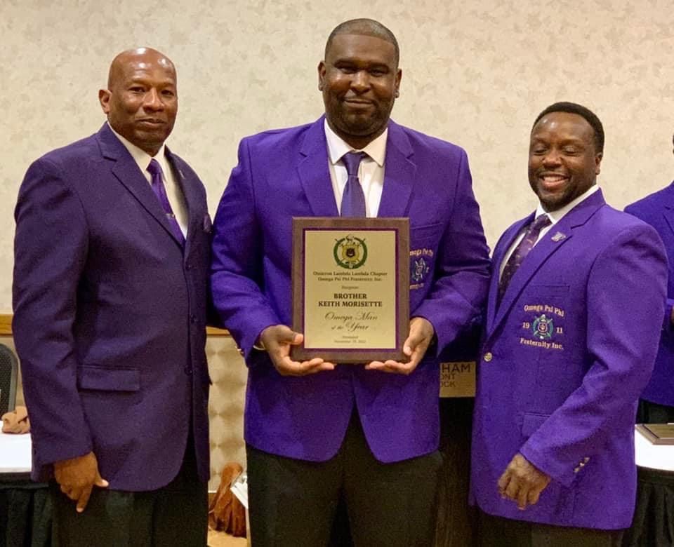 Three men in purple jackets holding an award plaque in a room with a table.