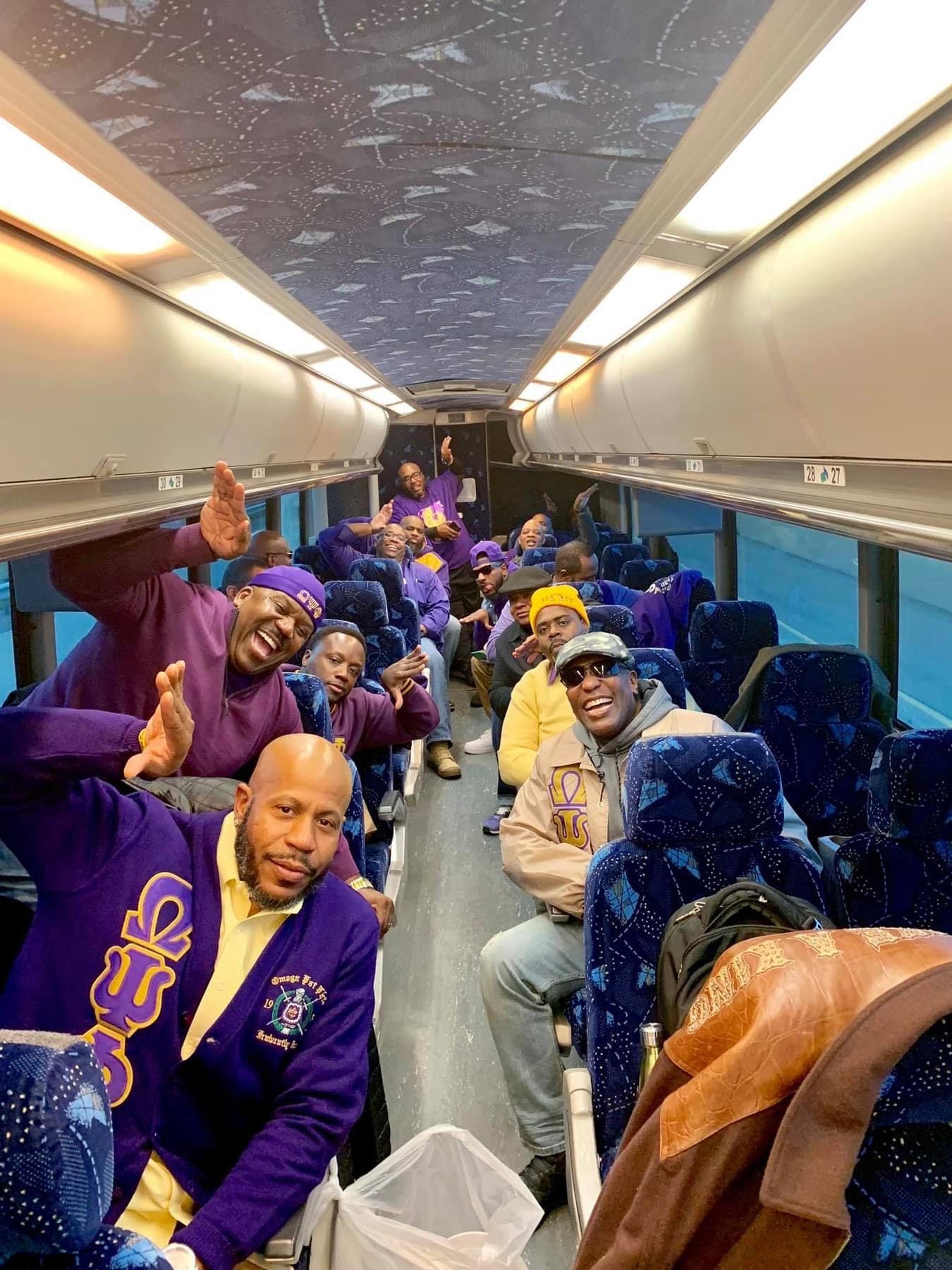People in a bus, many wearing purple, smiling and waving. Interior view.
