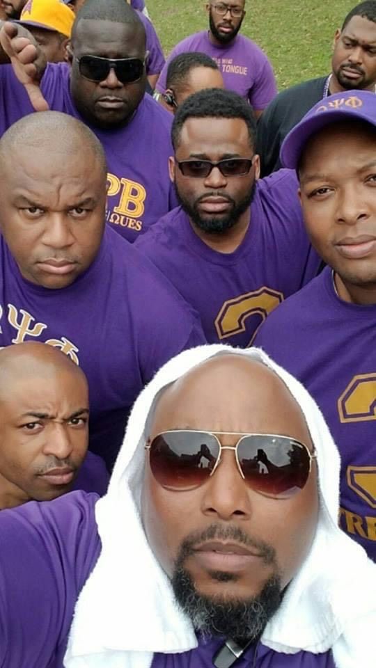 Group of men in purple shirts, possibly a fraternity, taking a selfie. Some wear sunglasses and have serious expressions.