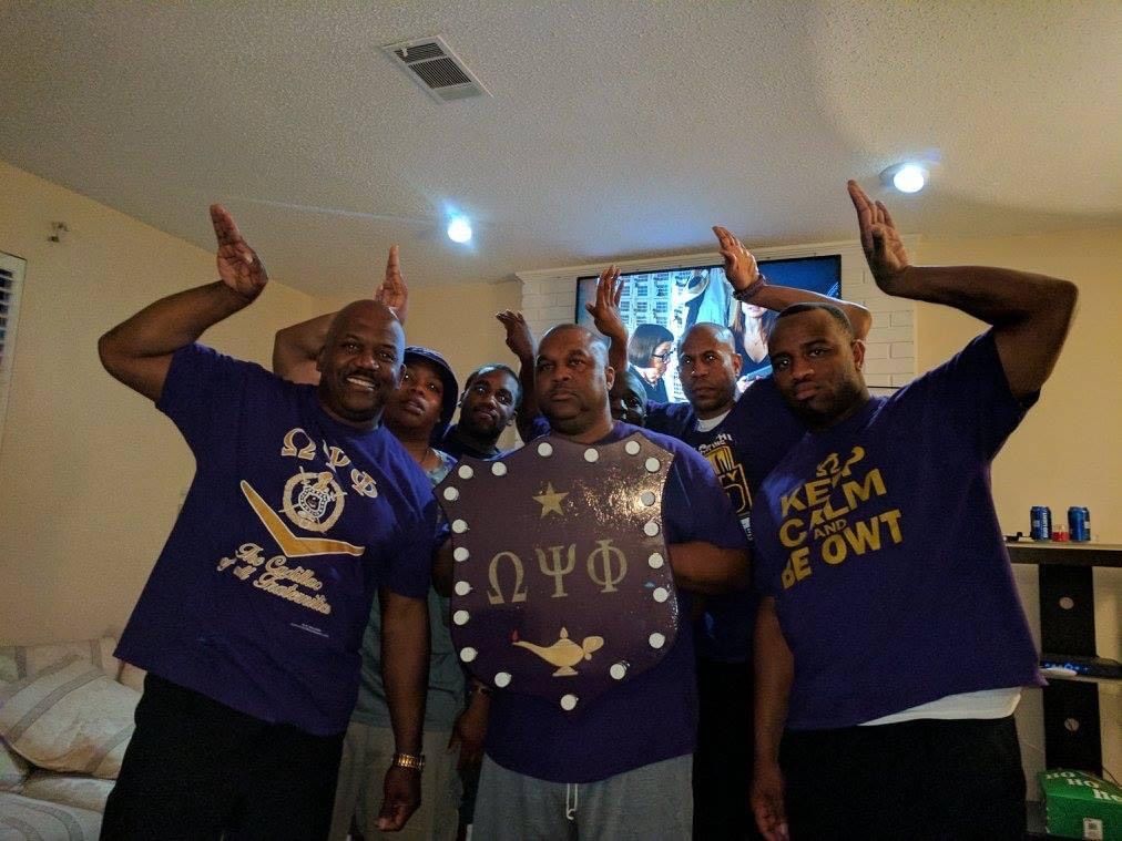 Group of men in purple shirts, some holding up hands, posing with a shield that has greek letters.