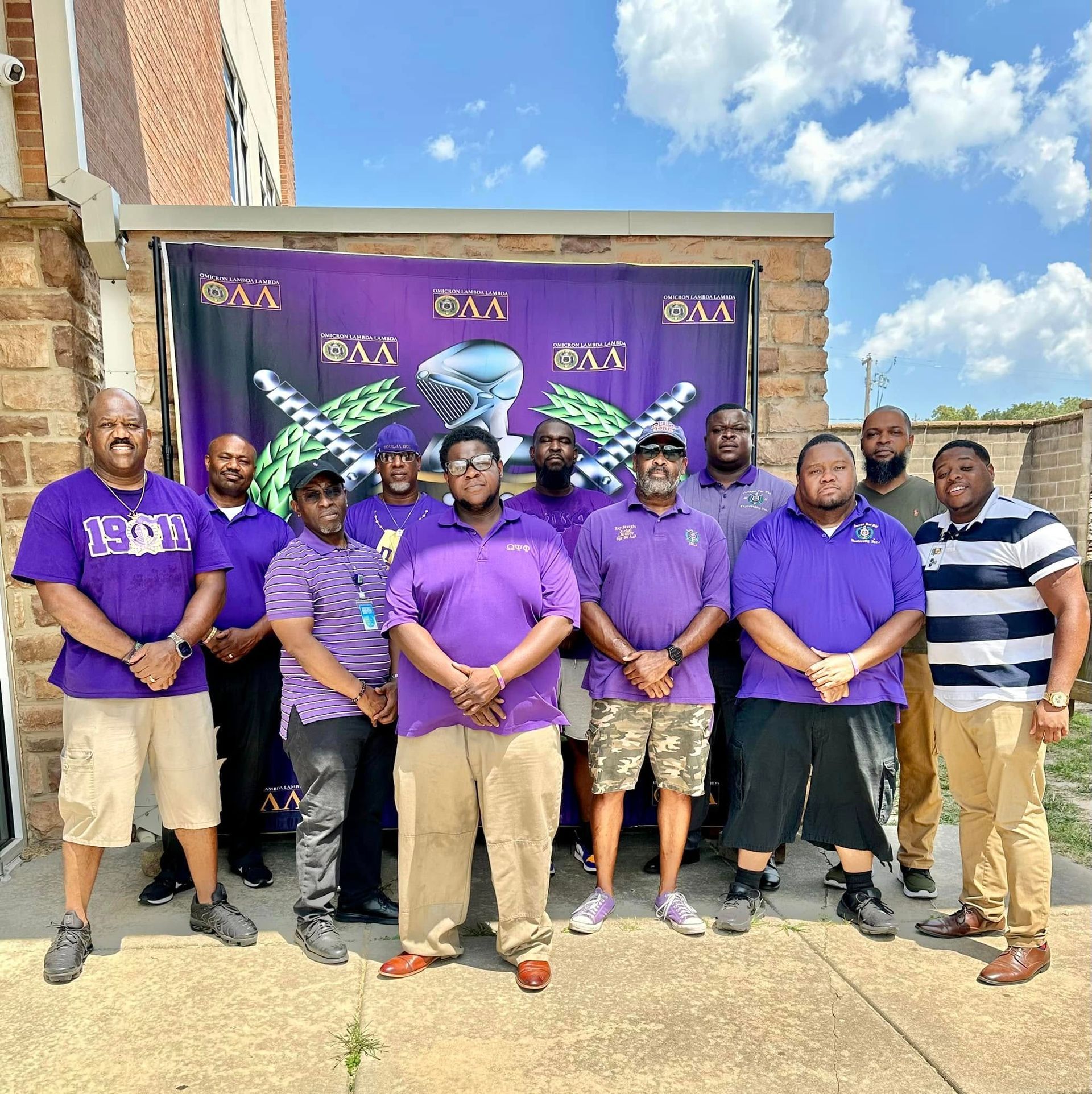 Group of men in purple attire pose in front of a banner. Outdoors, sunny day.