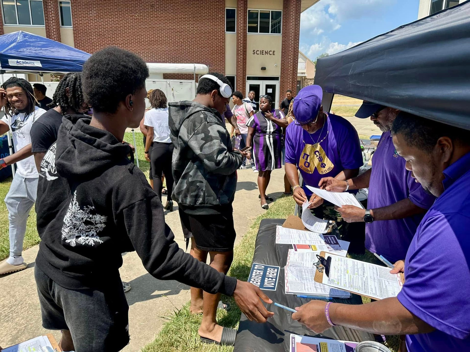 People at an outdoor event near a brick building, signing up at a table. Purple and black attire is prevalent.
