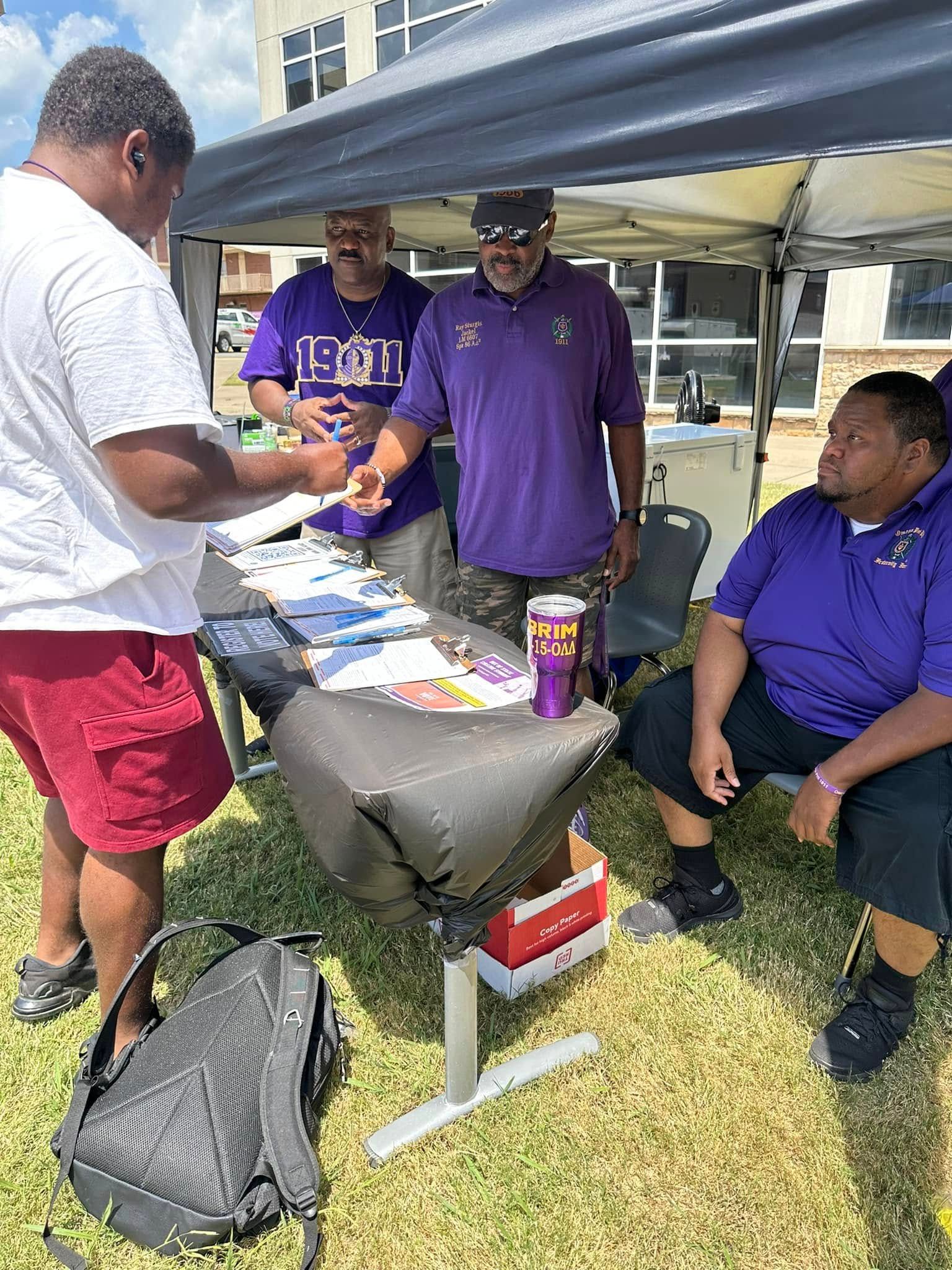 People at a table under a canopy, one hands a paper to another. Purple shirts, outdoors.
