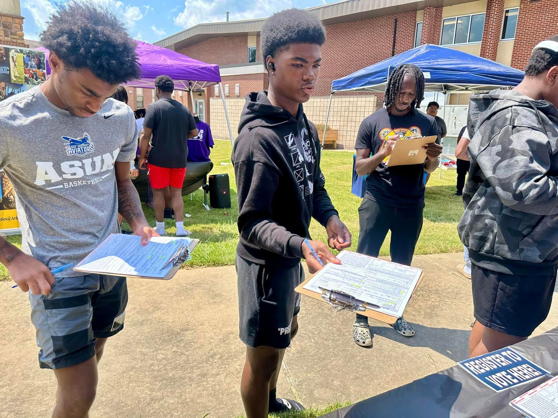 People at an outdoor table reviewing documents. Setting appears to be an event with tents, buildings, and grass.