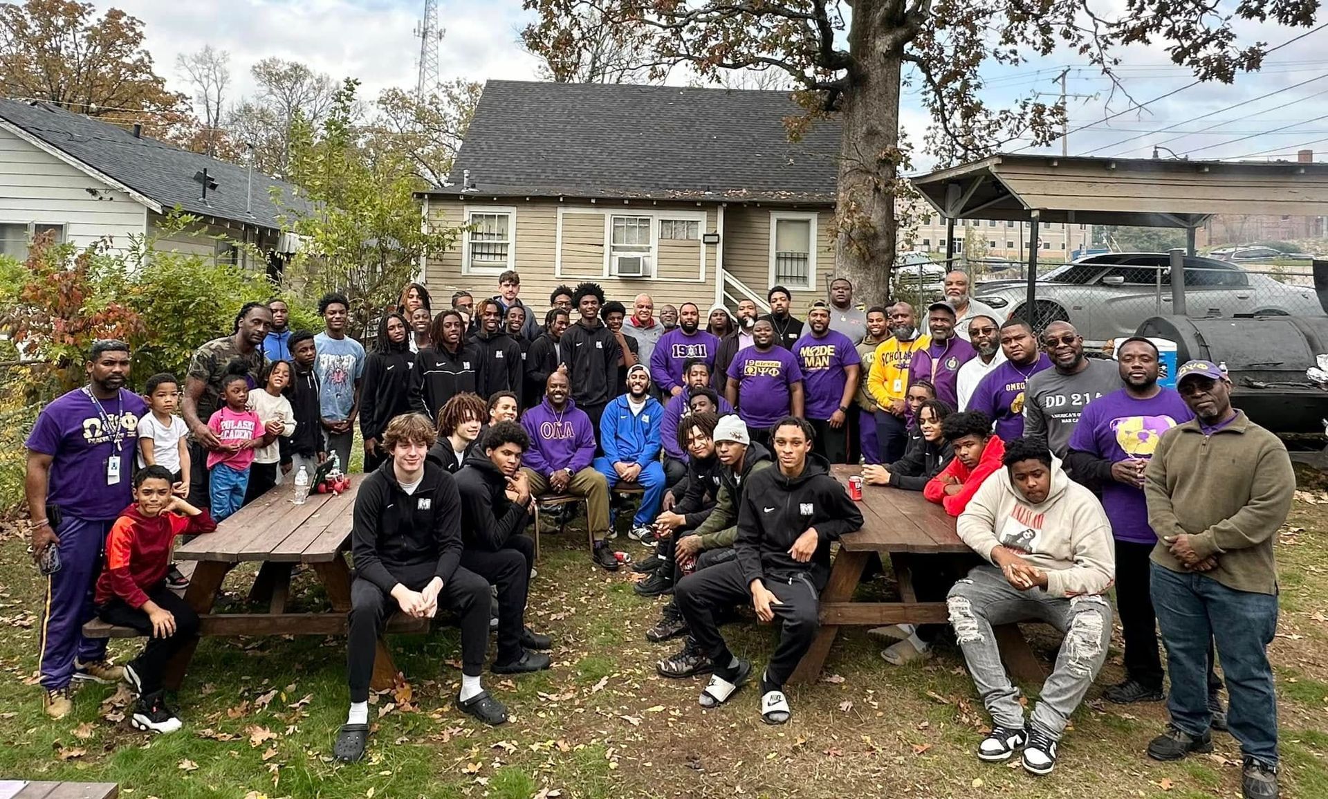 Large group of people gathered outdoors, some wearing purple, near houses.