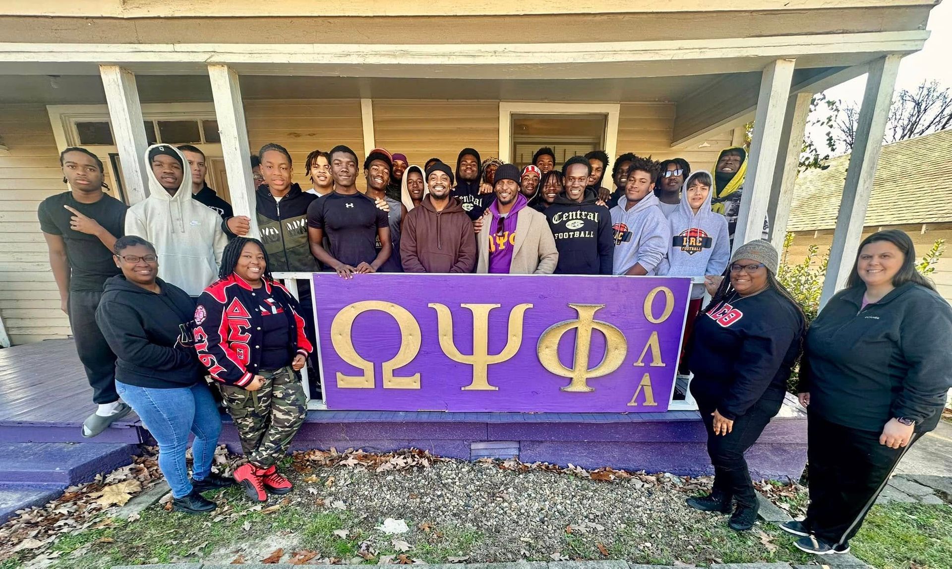 Group of people posing in front of a house, holding a purple sign with Greek letters.