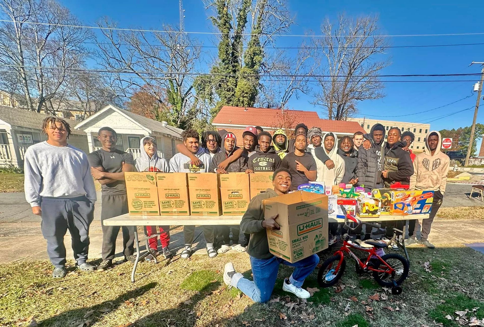 Group of people with donations and toys on a sunny day outdoors. Boxes of food are on a table.