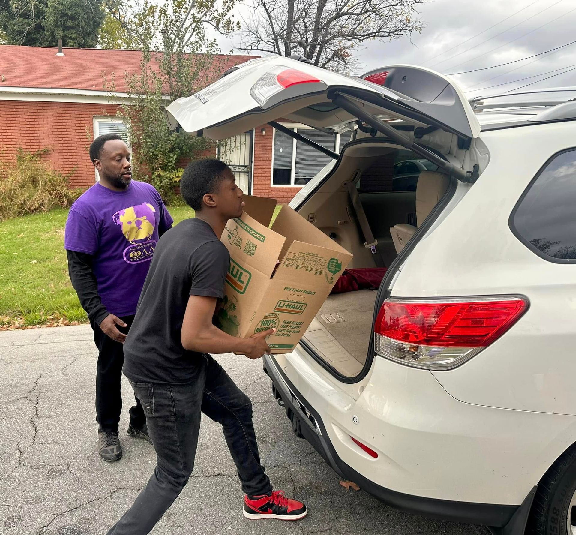 Man helping another load a box into a car trunk. Other man stands nearby. Cloudy day.