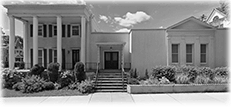 A grayscale photo of a stately building with a classical facade. It has large white columns and landscaped gardens.