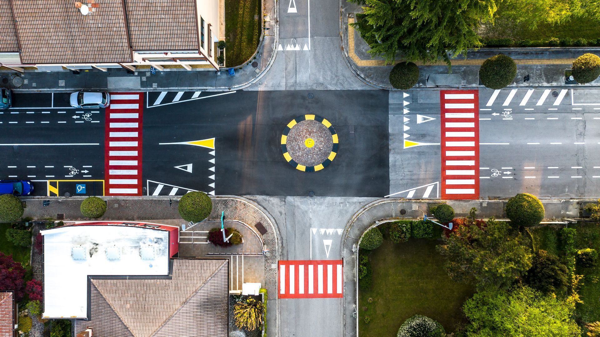 Vista dall'alto di un piccolo incrocio stradale con una rotonda dipinta, attraversamenti pedonali ed edifici residenziali.