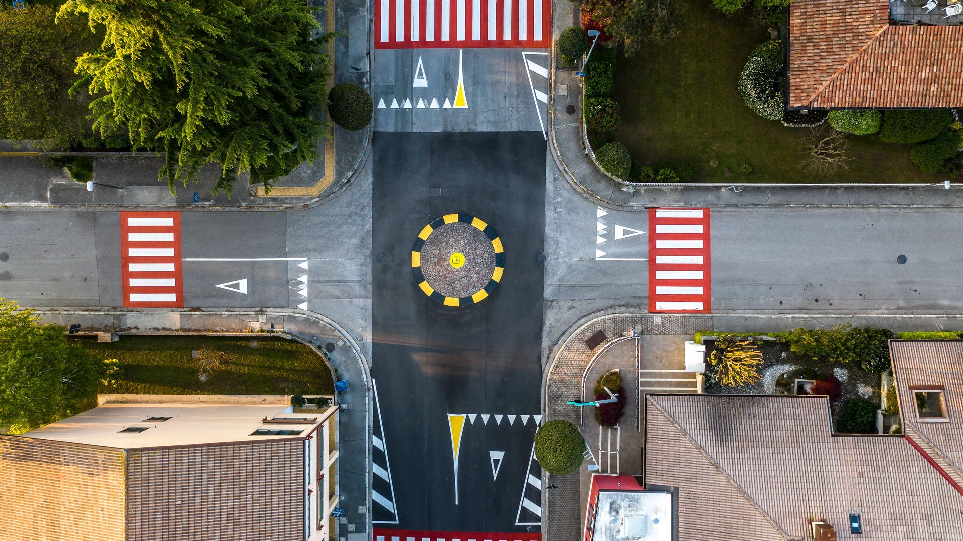 Vista dall'alto di un piccolo incrocio stradale con una rotonda dipinta, attraversamenti pedonali ed edifici residenziali.