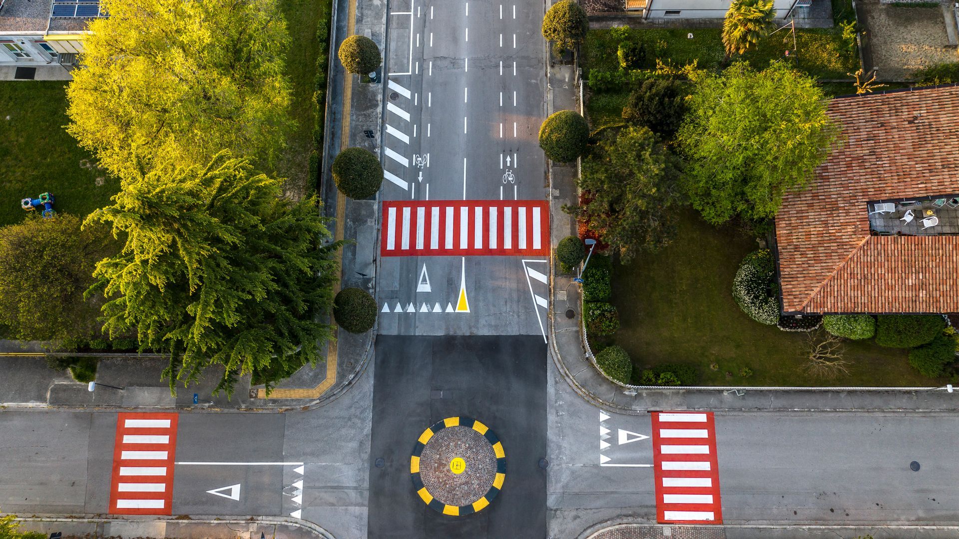 Vista aerea di un incrocio stradale con strisce pedonali dipinte di rosso e bianco e una rotonda con un disegno di fiori gialli.