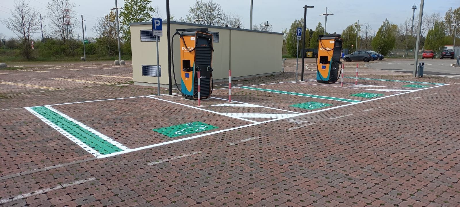 Stazione di ricarica per veicoli elettrici con due caricabatterie, posti auto dipinti su pavimentazione in mattoni e un piccolo edificio sullo sfondo.