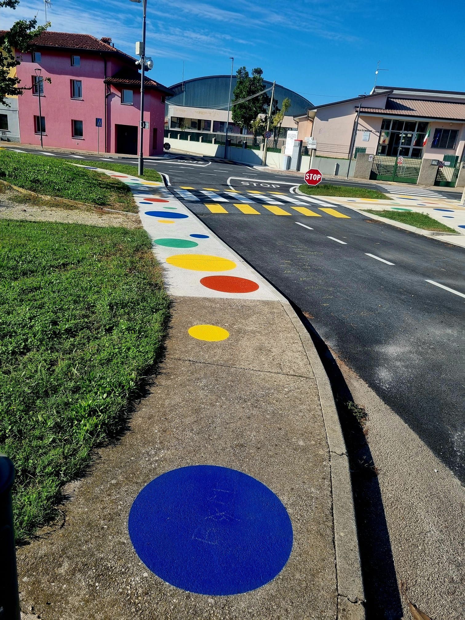 Scena di strada con un marciapiede colorato caratterizzato da grandi cerchi dipinti, un attraversamento pedonale ed edifici.