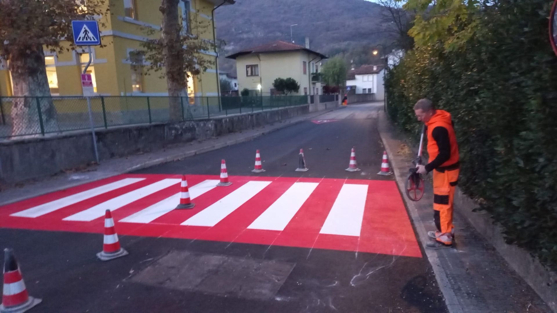 Un operaio ridipinge un attraversamento pedonale con strisce rosse e bianche. Sono visibili coni e segnali stradali.