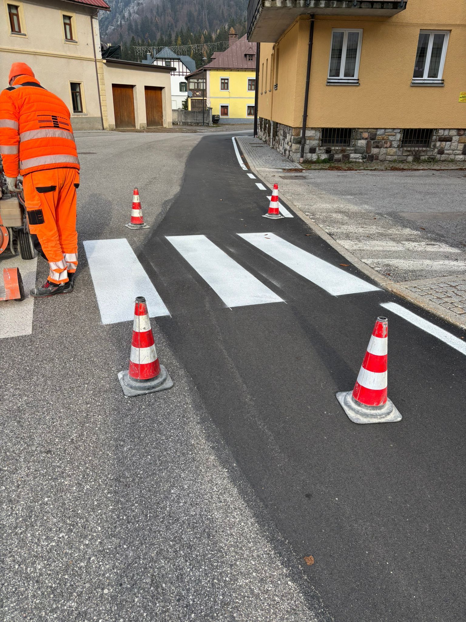 Operaio in tuta arancione che dipinge le strisce pedonali su una strada asfaltata; coni arancioni indicano la vernice fresca; edifici sullo sfondo.