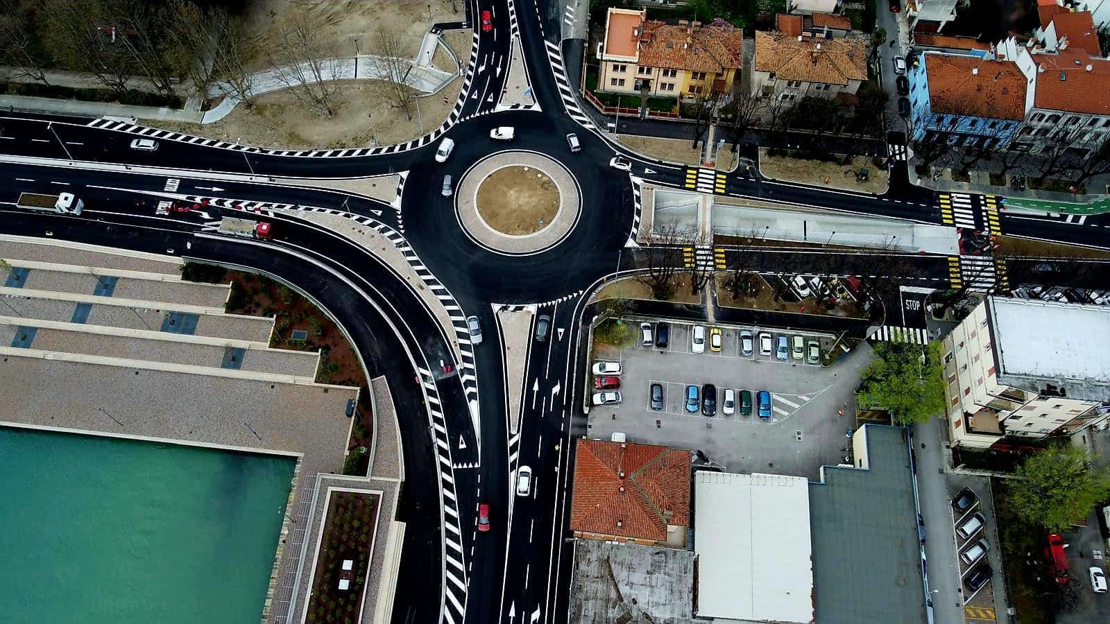 Vista aerea di una rotonda con traffico, edifici e un elemento d'acqua sulla sinistra.
