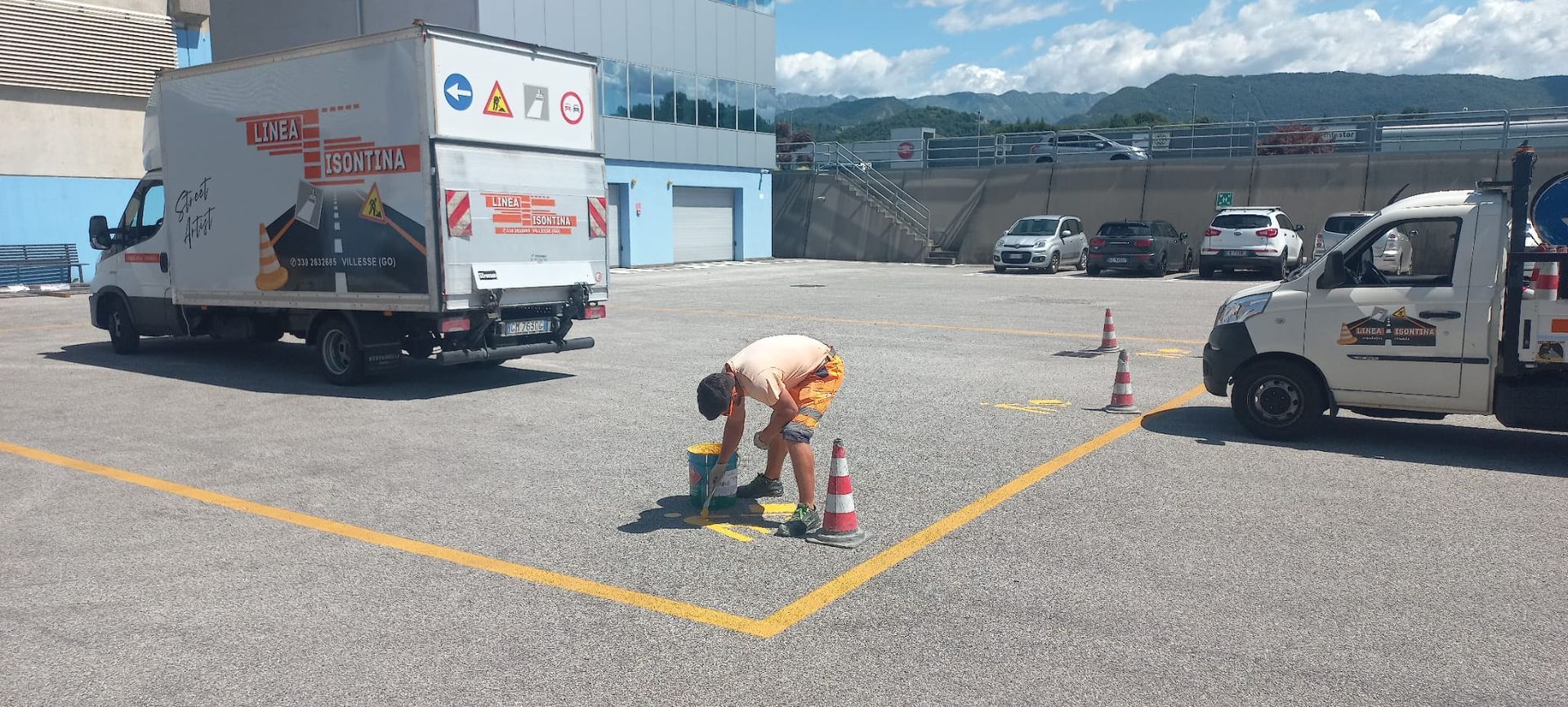 Una persona dipinge una linea gialla in un parcheggio, con due camion e delle auto sullo sfondo.