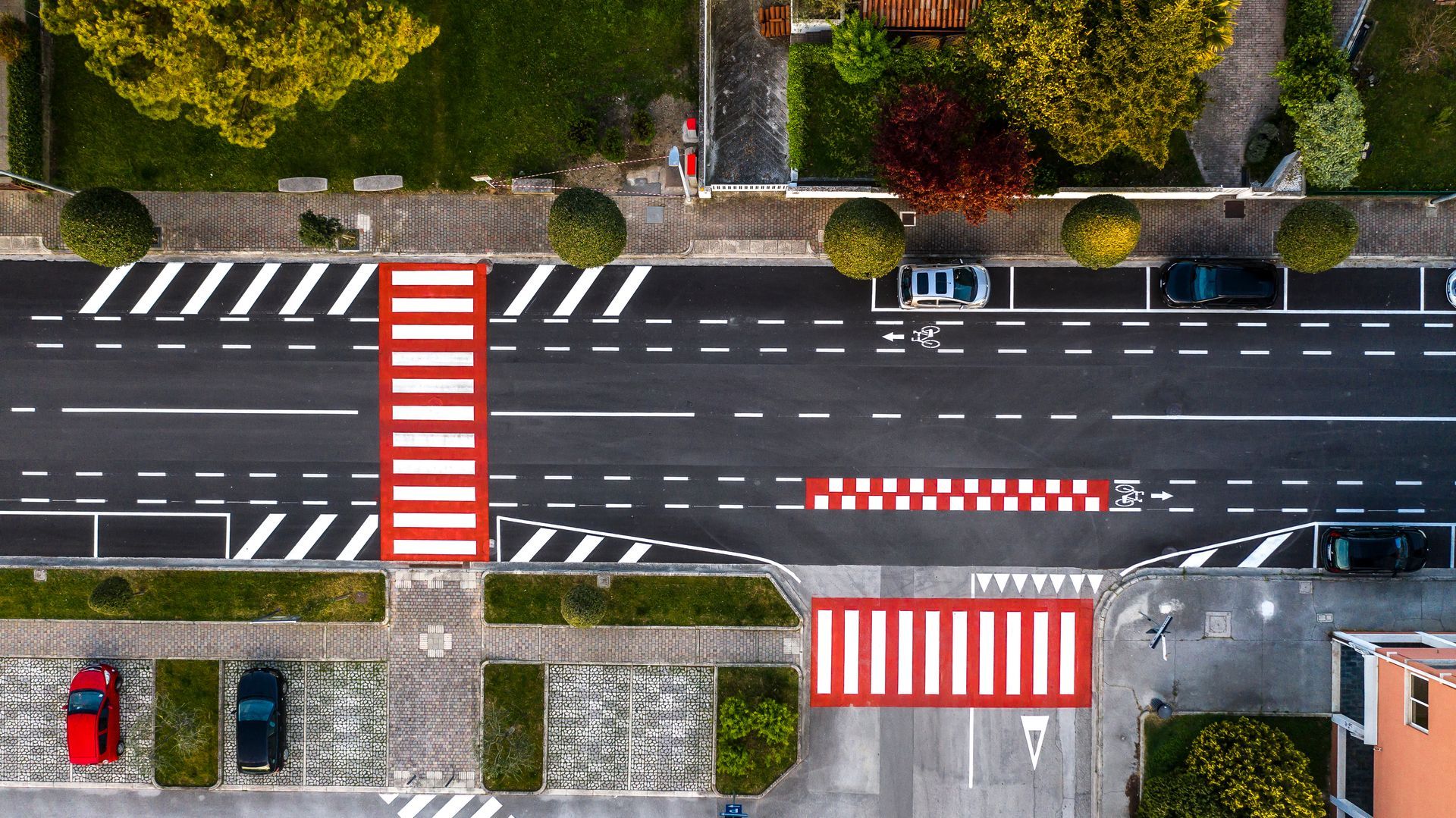 Vista dall'alto di una strada cittadina con attraversamenti pedonali, segnaletica orizzontale a strisce e auto parcheggiate.