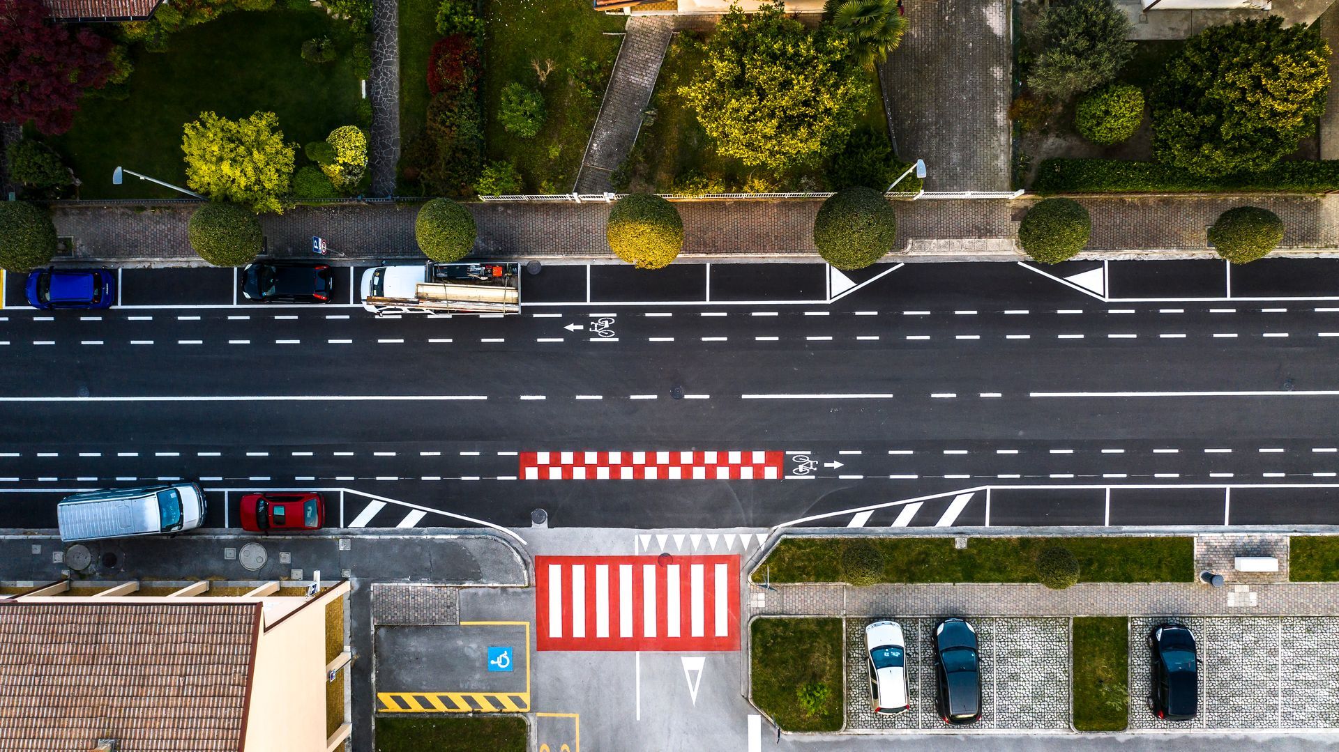 Vista dall'alto di una strada con auto parcheggiate su entrambi i lati, un attraversamento pedonale e alberi lungo i marciapiedi.