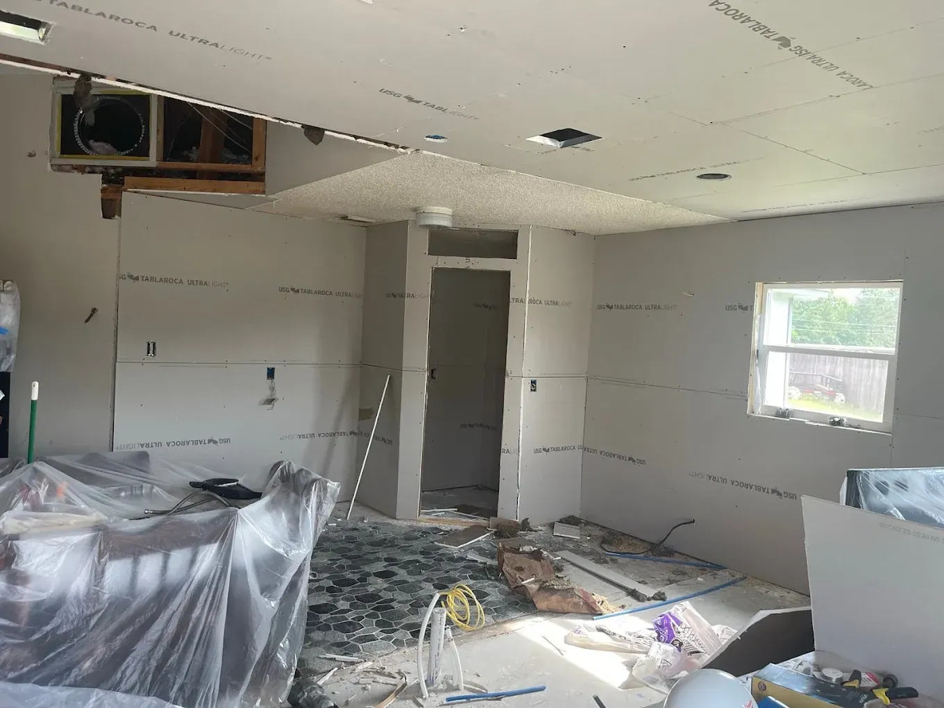 Interior of a room under construction, drywall installed on walls and ceiling. Debris and tools litter the floor.